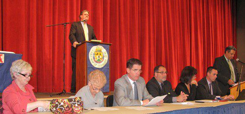 TOWN MODERATOR William Carroll stands on stage during last night’s Town Meeting while (from left) selectmen Betsy Sheeran, Phyllis Hull, Patrick Glynn, Paul DiNocco, Ann Santos and Brian Falvey are seated in foreground. Town Administrator Stephen P. Maio speaks at right. (Mark Sardella Photo)