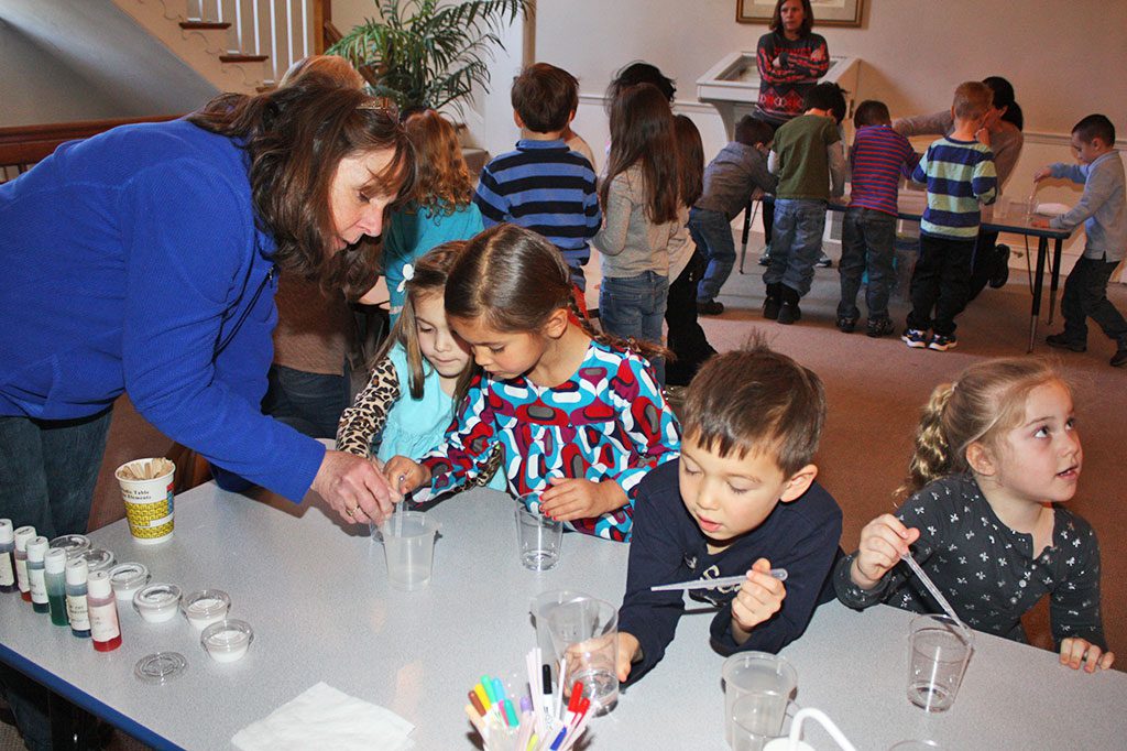 CHILDREN learn by doing and these 4-year-olds at Tower Day School were eager to learn how to squeeze the right number of water drops into a cup, the first step in making their own colorful slime, with the help of teacher Nancy Miller during the chemistry segment of Science Week. (Maureen Doherty Photo)